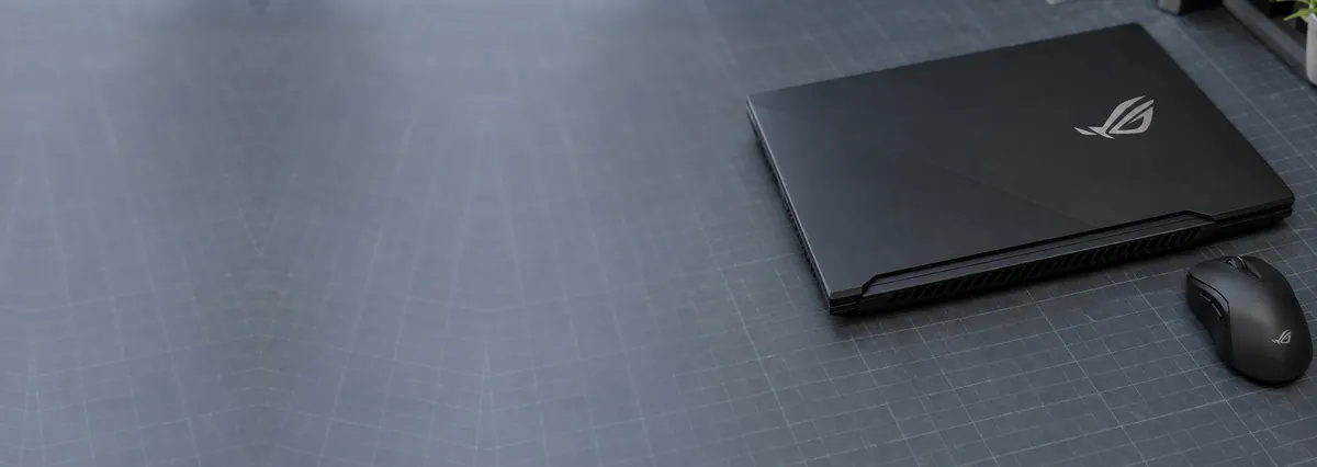 A closed black ASUS ROG gaming laptop beside a gaming mouse on a slate-grey workbench