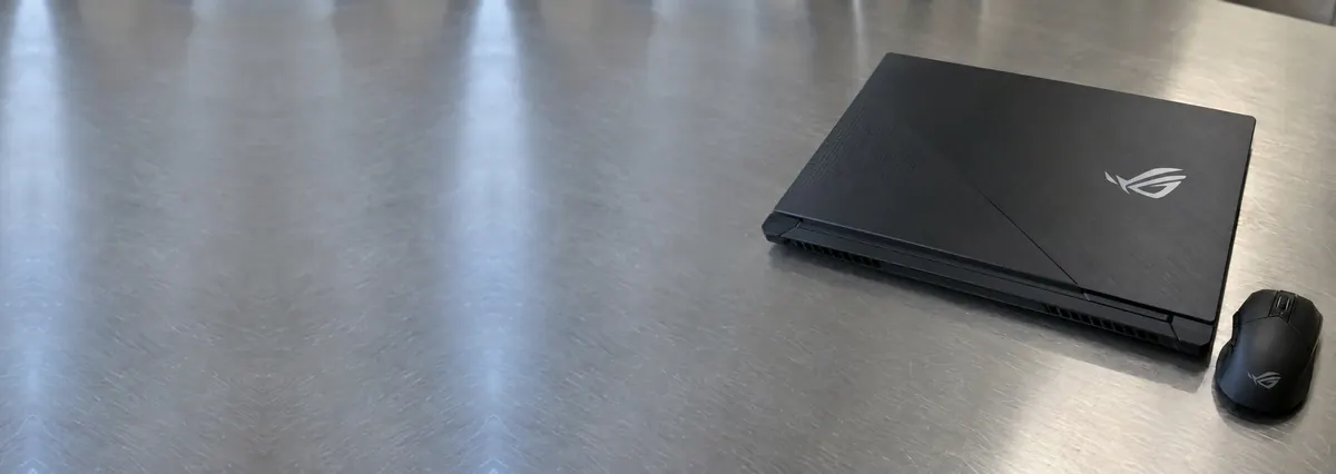 A closed black ASUS ROG gaming laptop beside a gaming mouse on a steel workbench