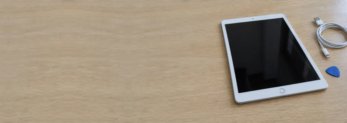 An Apple iPad lying face-up beside a charging cable and an opening pick on an oak desk top