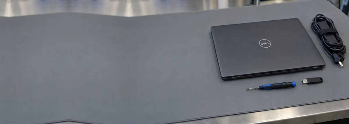 A closed charcoal Dell laptop beside a screwdriver, a flash drive and a charger cable on a grey anti-static mat