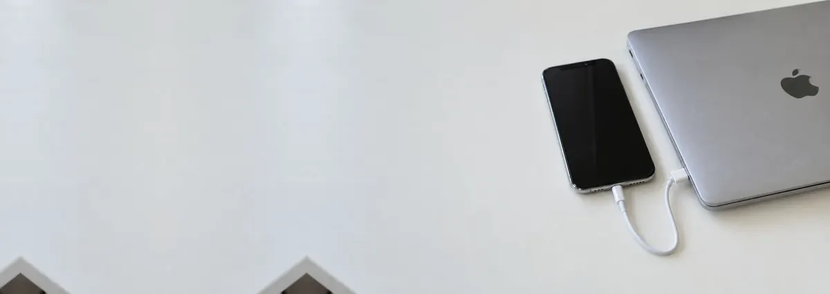An Apple iPhone connected by a cable to a closed MacBook on a white bench