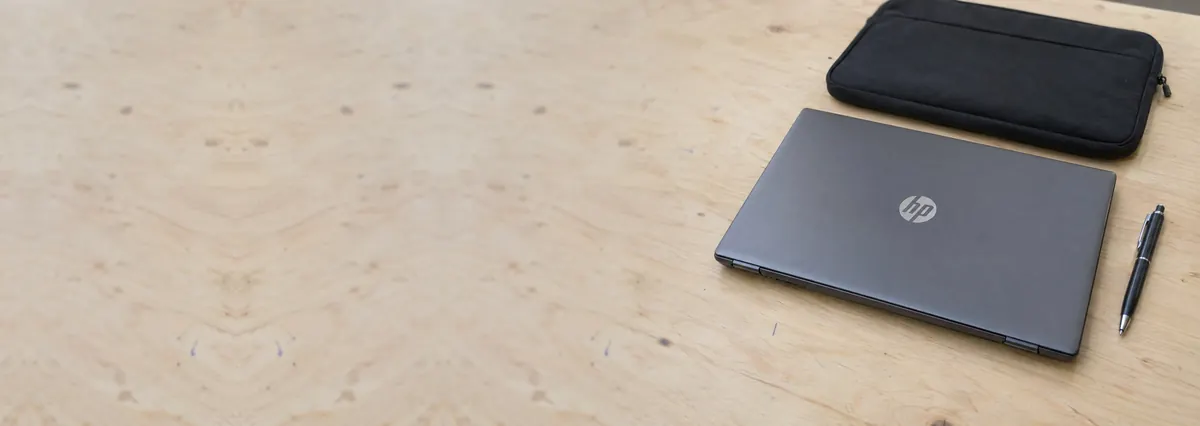 A closed dark-silver HP laptop beside a laptop sleeve and a pen on a plywood bench