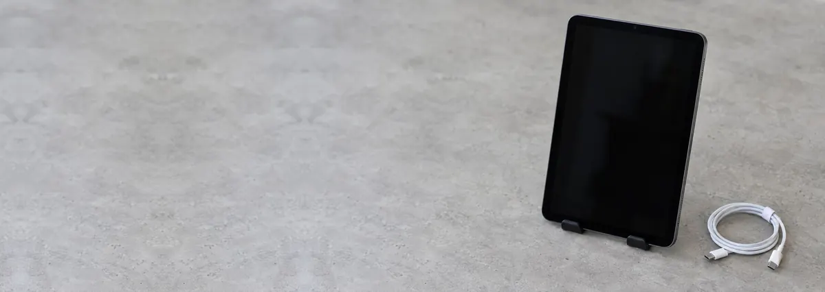 A space-grey Apple iPad on a stand beside a coiled charging cable on a concrete-effect workbench