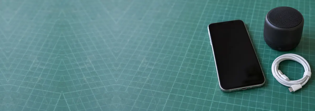 An Apple iPhone beside a small black speaker and a coiled cable on a green cutting mat
