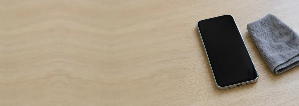 An Apple iPhone with its screen off beside a microfibre cloth on an oak desk top