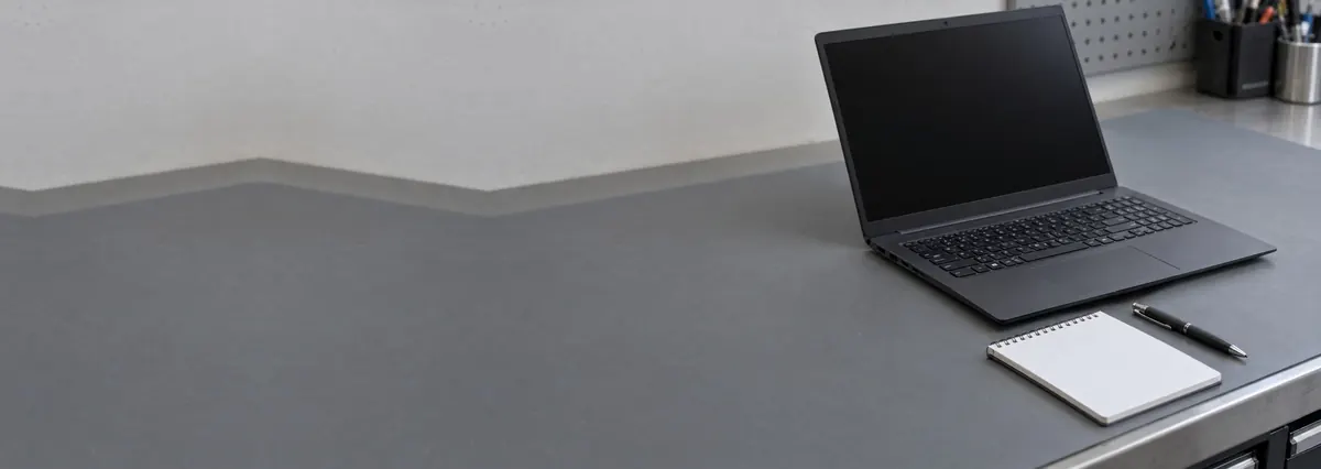 An open dark laptop with its screen off beside a notepad and a pen on a grey anti-static mat