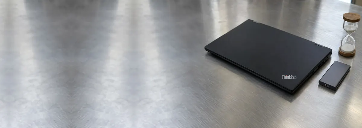 A closed Lenovo ThinkPad beside a slim SSD and a small hourglass on a steel workbench