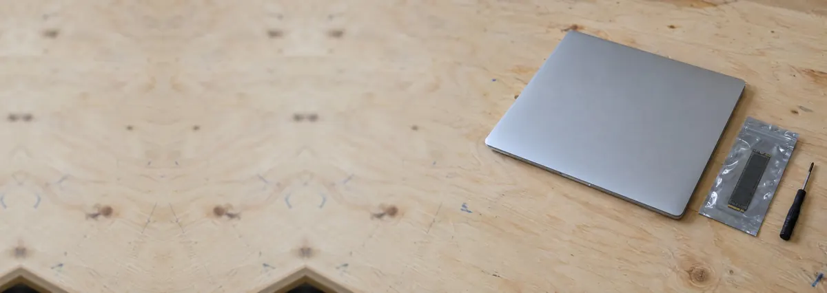 A closed laptop beside an SSD in an antistatic bag and a screwdriver on a plywood bench