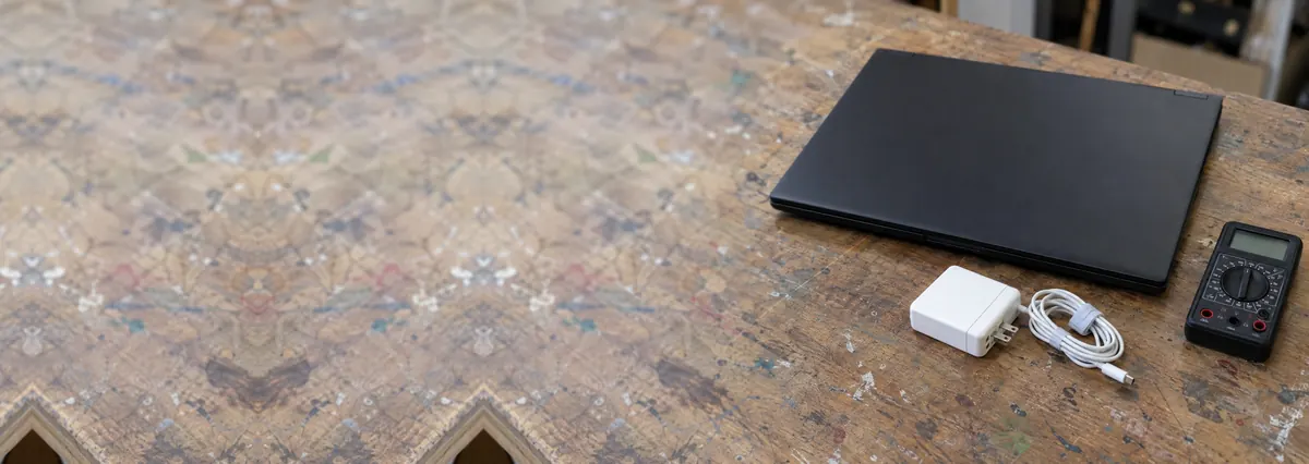 A closed black laptop beside an unplugged charger and a multimeter on a wooden bench
