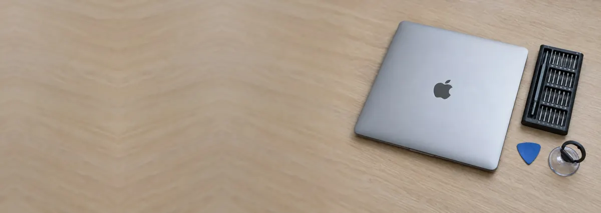 A closed Apple MacBook beside a screwdriver set, an opening pick and a suction handle on an oak desk top