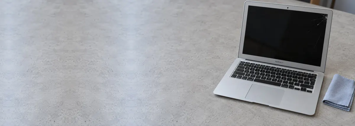 An open Apple MacBook with a cracked screen beside a lint-free cloth on a concrete-effect workbench