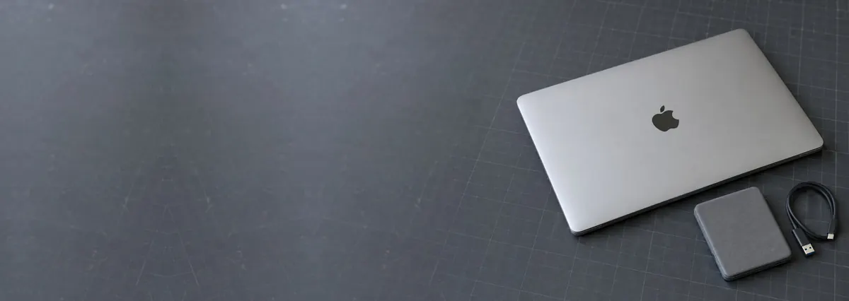 A closed Apple MacBook beside an external hard drive and a USB cable on a slate-grey workbench