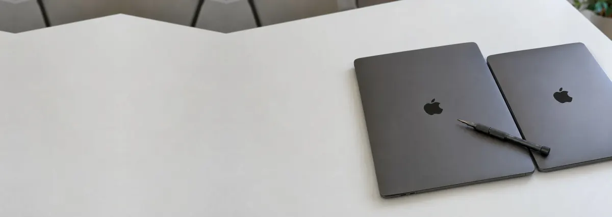 Two closed space-black Apple MacBook Pro laptops side by side with a screwdriver across them on a white bench