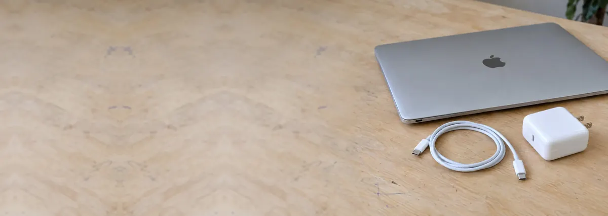 A closed Apple MacBook with an unplugged USB-C cable and a charging brick on a plywood bench
