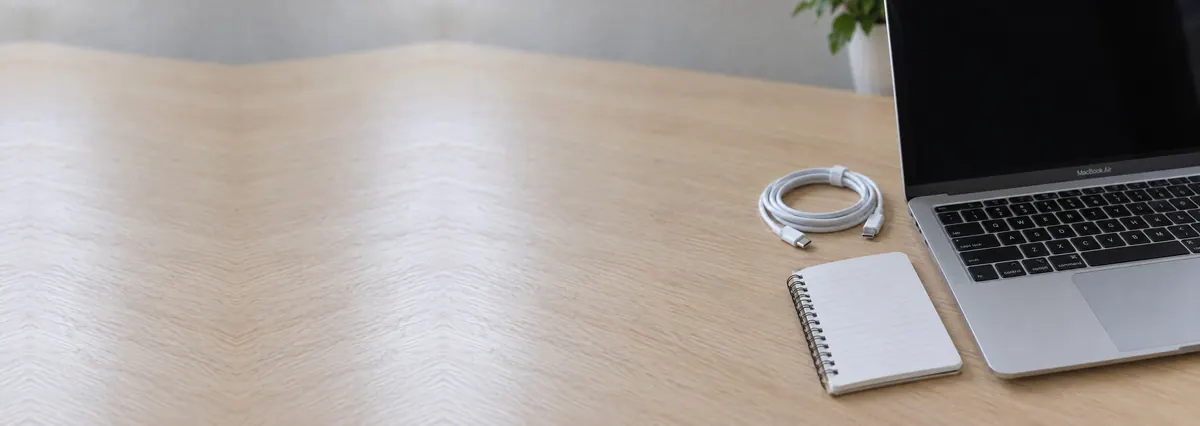 An open Apple MacBook with its screen off beside a coiled cable and a notepad on an oak desk top