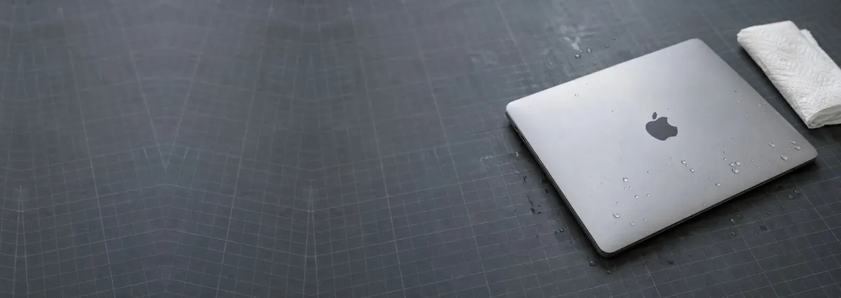 A closed Apple MacBook with water droplets beside a paper towel on a slate-grey workbench