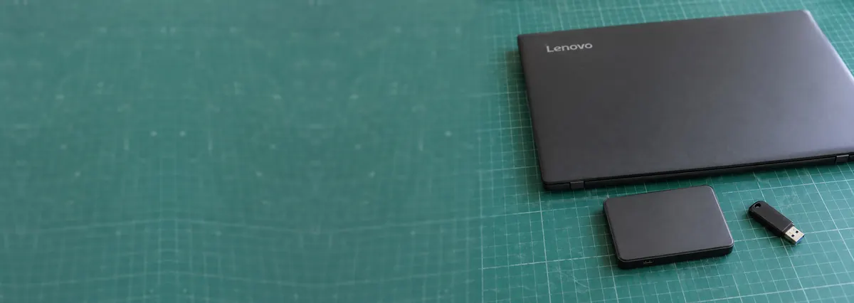A closed Lenovo laptop beside an external hard drive and a flash drive on a green cutting mat