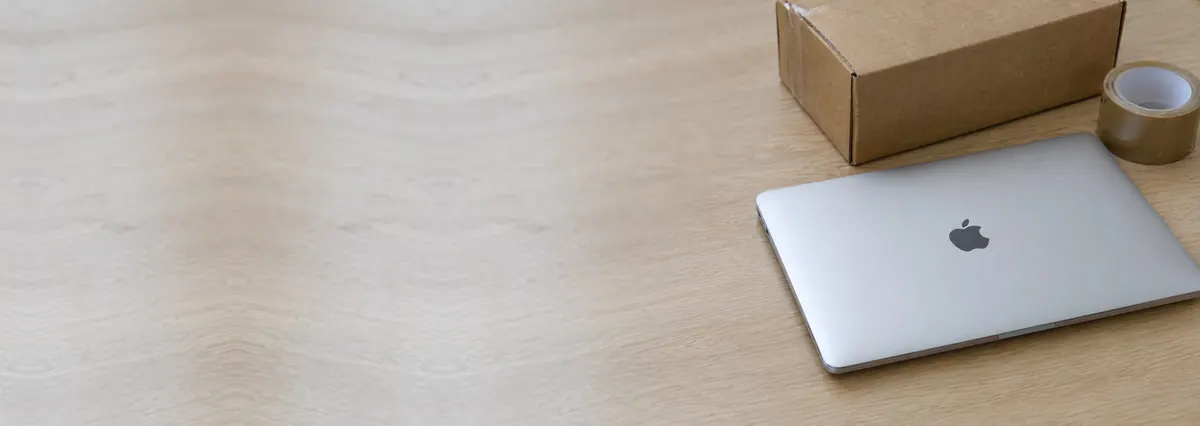 A closed Apple MacBook beside a cardboard shipping box and a roll of tape on an oak desk top