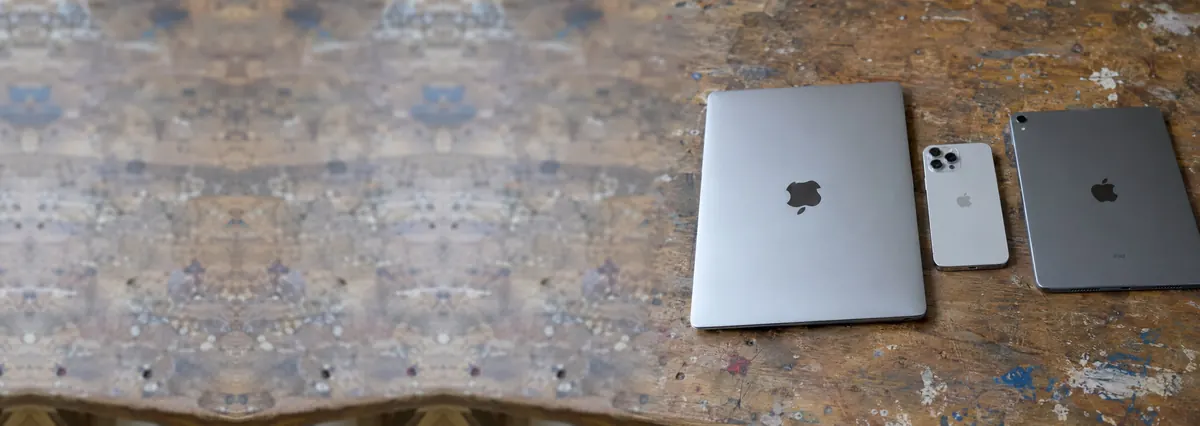 A closed Apple MacBook, an iPhone and an iPad side by side on a wooden bench