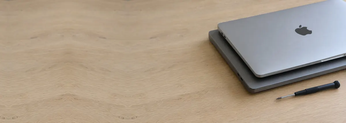 Two closed Apple MacBooks, a silver one and a space-grey one, beside a screwdriver on an oak desk top