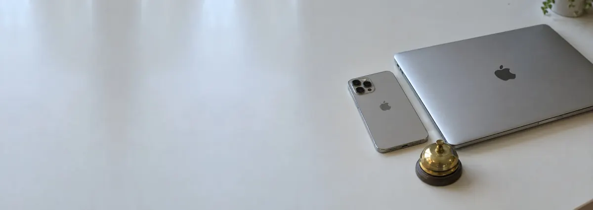 A closed Apple MacBook and an iPhone beside a small brass bell on a white bench