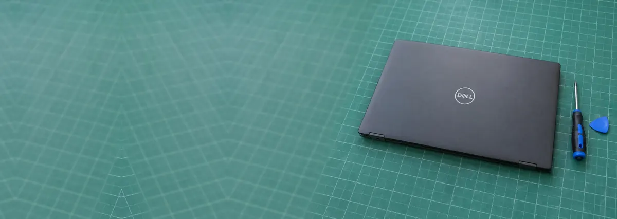 A closed dark-grey business Dell laptop on a green cutting mat beside a screwdriver and an opening pick
