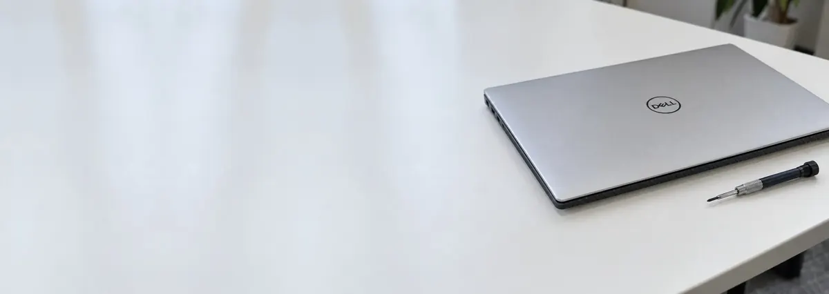 A closed silver thin Dell laptop with a carbon-fibre base edge beside a precision screwdriver on a white bench