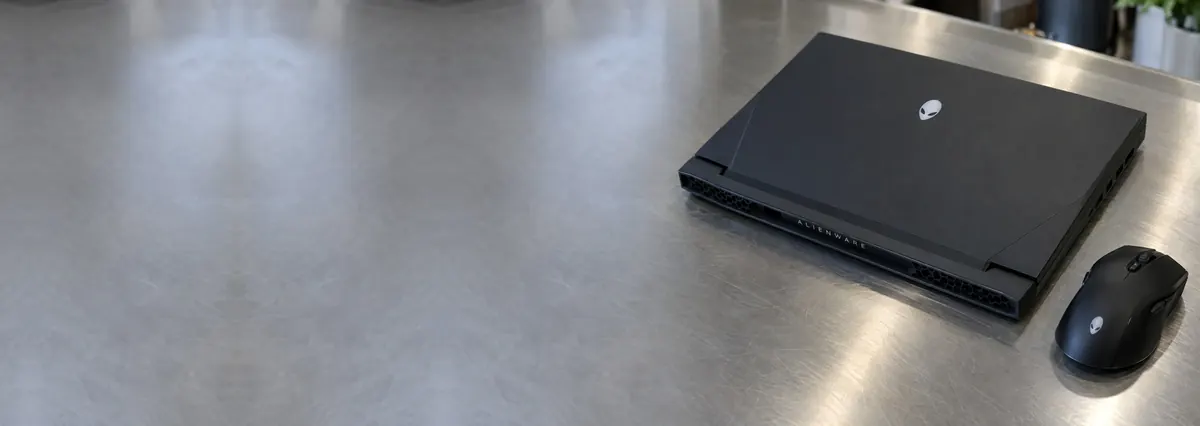 A closed black Alienware gaming laptop beside a gaming mouse on a steel workbench