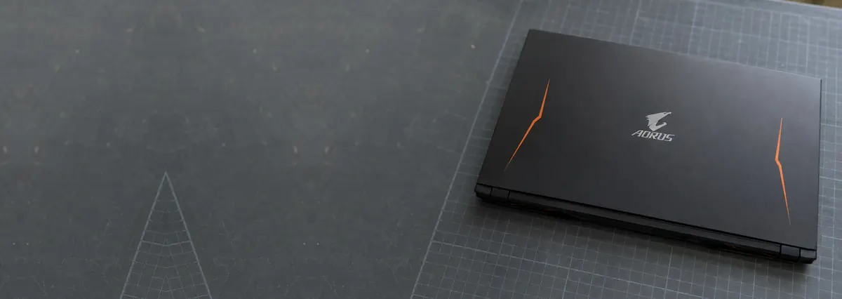 A closed matte-black Gigabyte AORUS laptop with orange accents on a slate-grey workbench