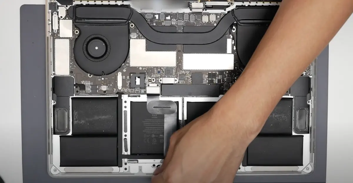 Inside an opened MacBook, with a technician's hand lifting the battery pull tab