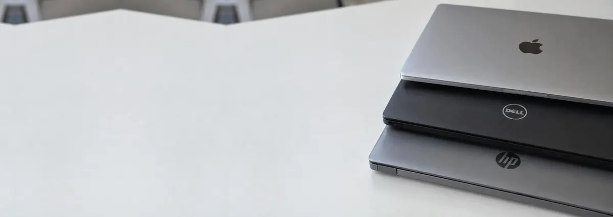 Three closed laptops, an Apple MacBook, a Dell and an HP, stacked on a white bench