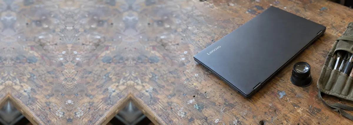 A closed Lenovo laptop beside a magnifying loupe and a toolkit roll on a wooden bench