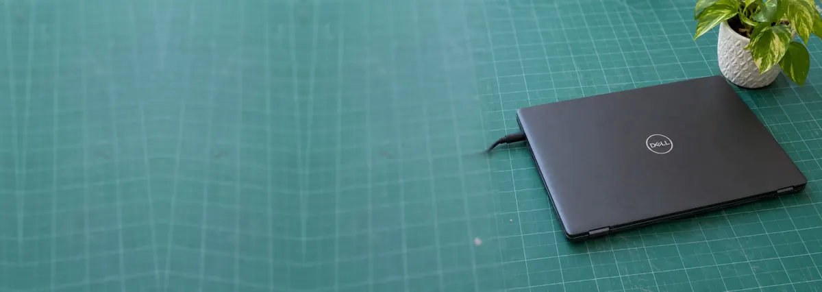 A closed Dell laptop with a charger plugged in beside a small plant on a green cutting mat