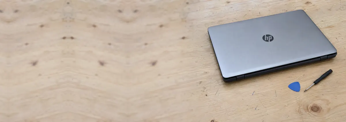 A closed silver-and-black HP laptop on a pale plywood bench beside an opening pick and a screwdriver