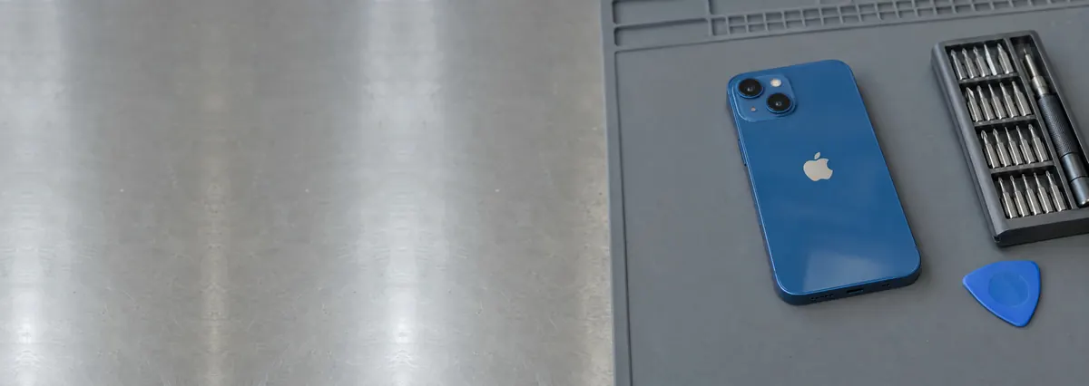 A blue Apple iPhone lying face-down on a grey anti-static mat beside a screwdriver set and an opening pick