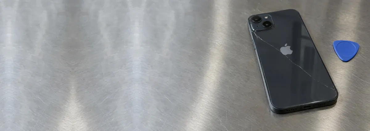 An Apple iPhone with a cracked glass back beside an opening pick on a steel workbench
