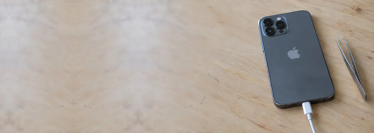 An Apple iPhone with a charging cable plugged into its port beside fine tweezers on a plywood bench