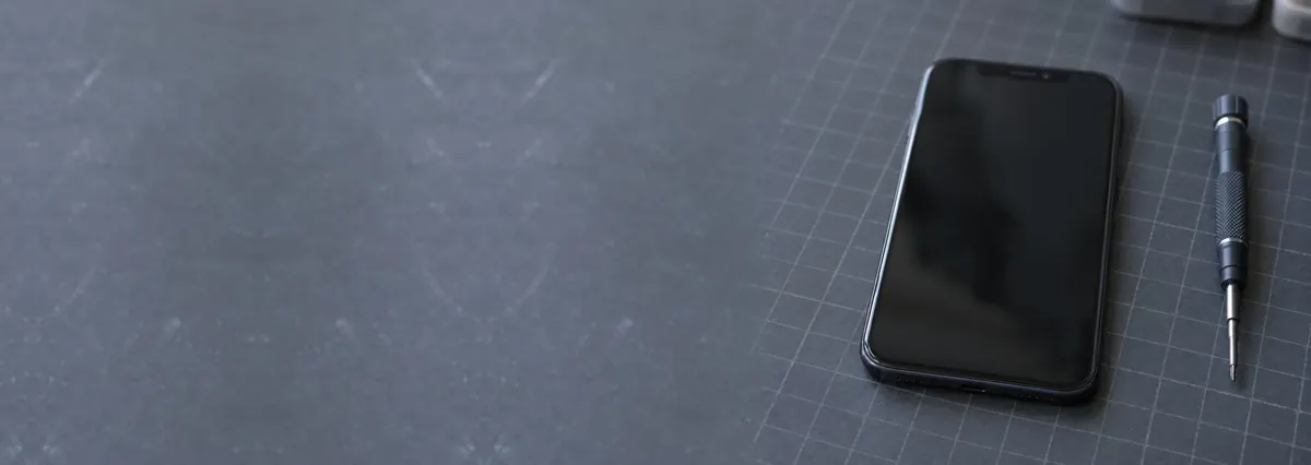 A smartphone lying face-up with its screen off beside a fine screwdriver on a slate-grey workbench