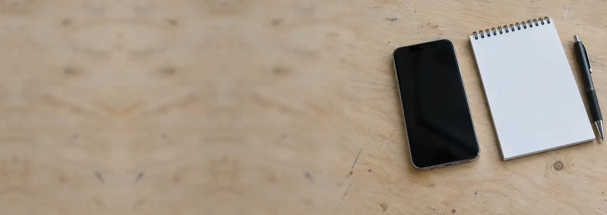 An Apple iPhone beside a notepad and a pen on a plywood bench
