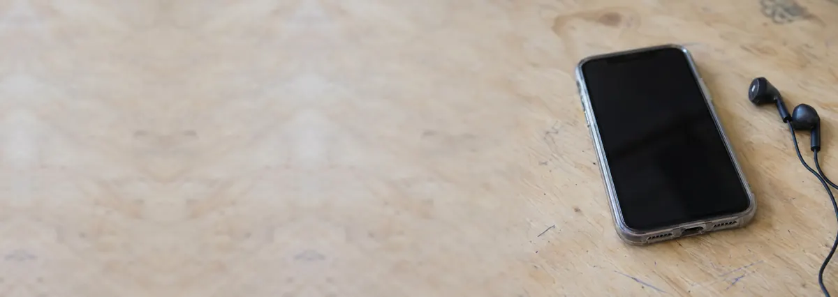 A smartphone lying face-up with its screen off beside wired earbuds on a plywood bench