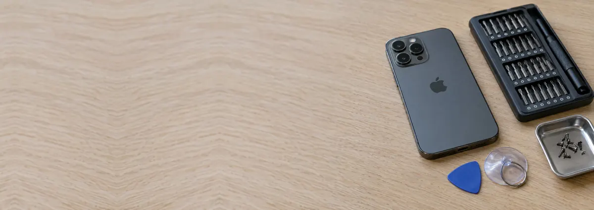 An Apple iPhone Pro lying face-down beside a screwdriver set, an opening pick and a suction cup on an oak desk top