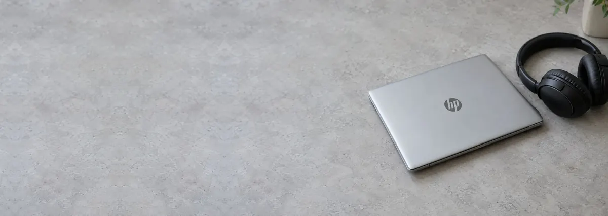 A closed silver HP laptop beside black over-ear headphones on a concrete-effect workbench
