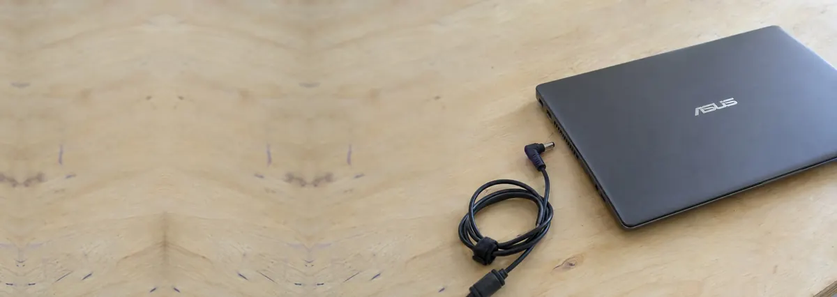 A closed ASUS laptop with an unplugged charger cable on a plywood bench