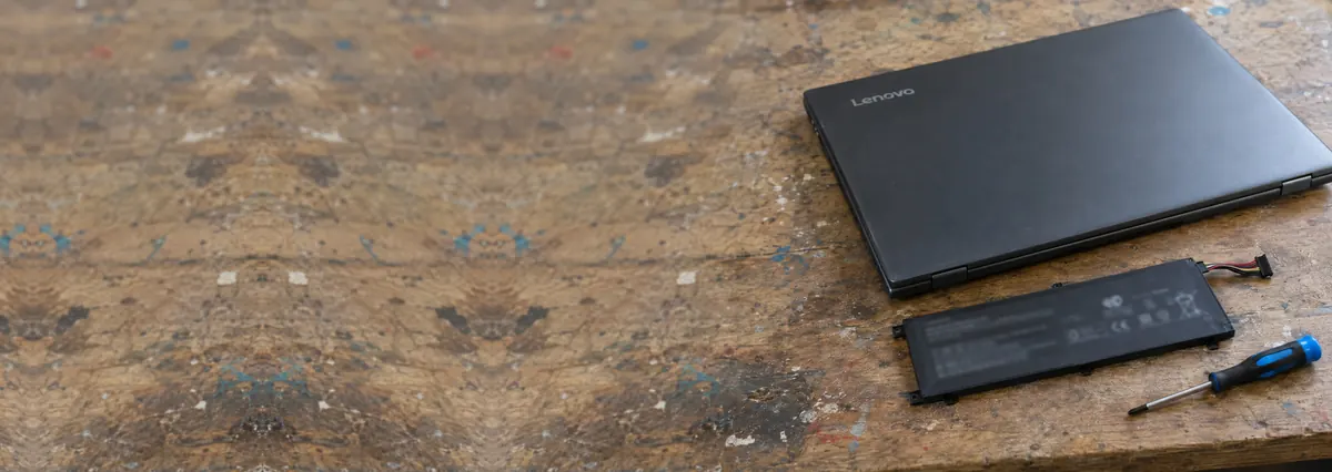 A closed Lenovo laptop beside a flat black battery pack and a screwdriver on a wooden bench
