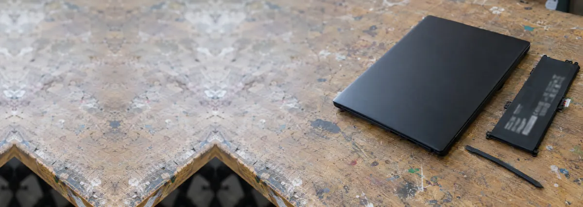 A closed black laptop beside a flat battery pack and a spudger on a wooden bench