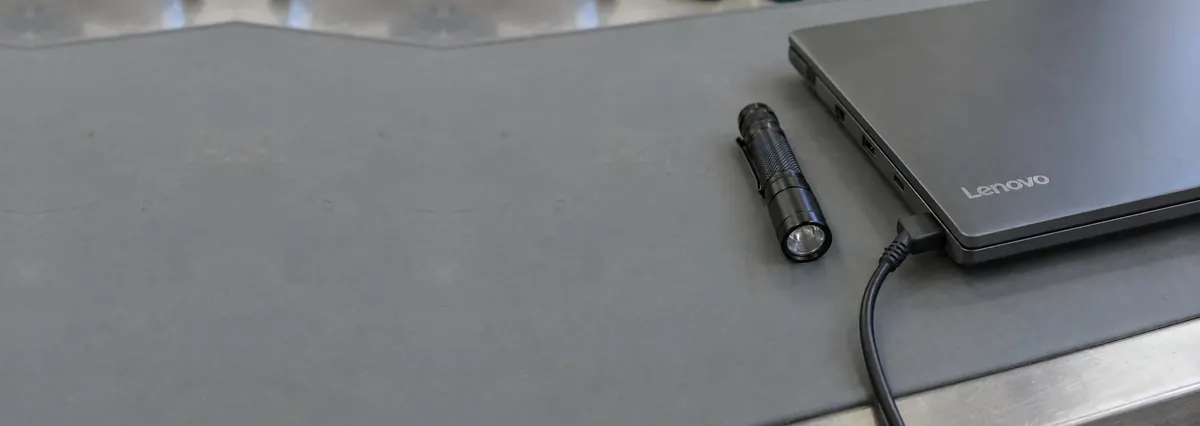 A closed Lenovo laptop with a charger cable plugged into its side port on a grey anti-static mat