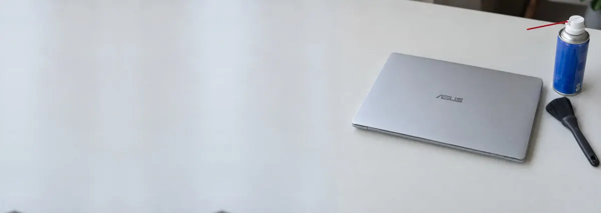 A silver ASUS laptop lying closed on a white bench beside a can of compressed air and a soft brush