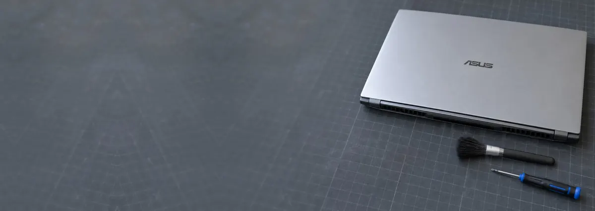 A closed silver ASUS laptop seen from its vent side beside a brush and a screwdriver on a slate-grey workbench