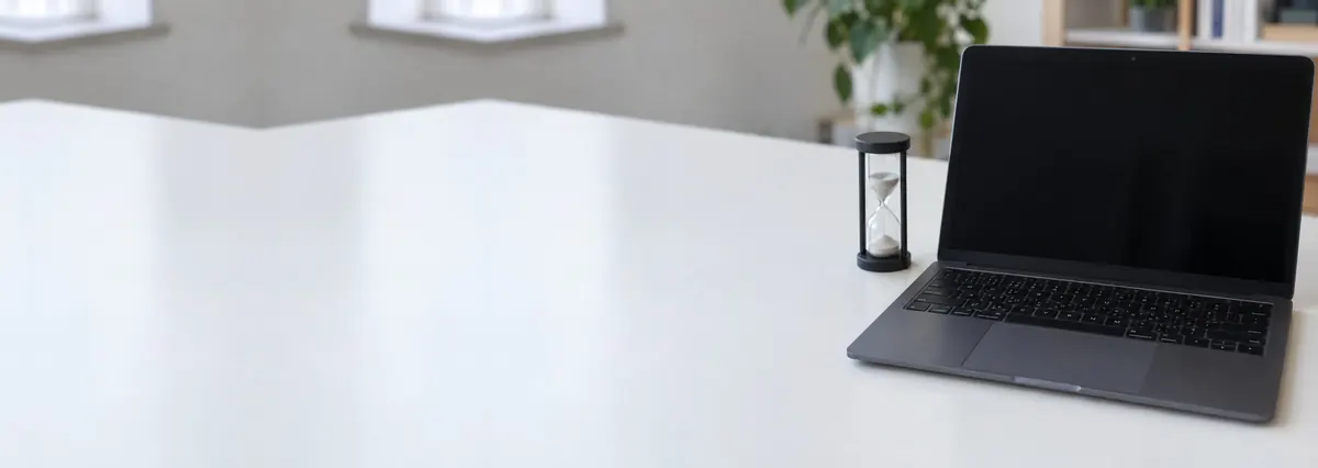 An open dark laptop with its screen off beside a small sand timer on a white bench