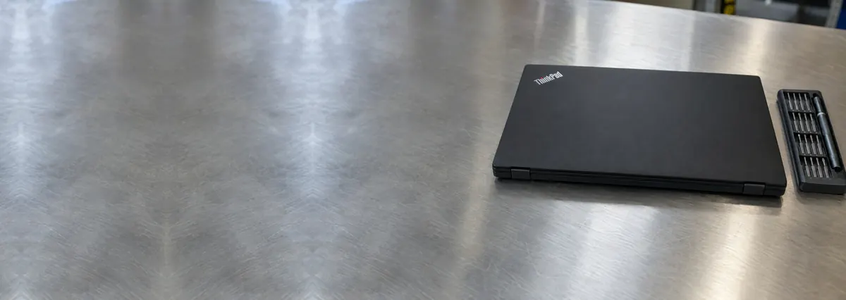 A closed black Lenovo ThinkPad laptop seen from its hinge side beside a screwdriver set on a steel workbench